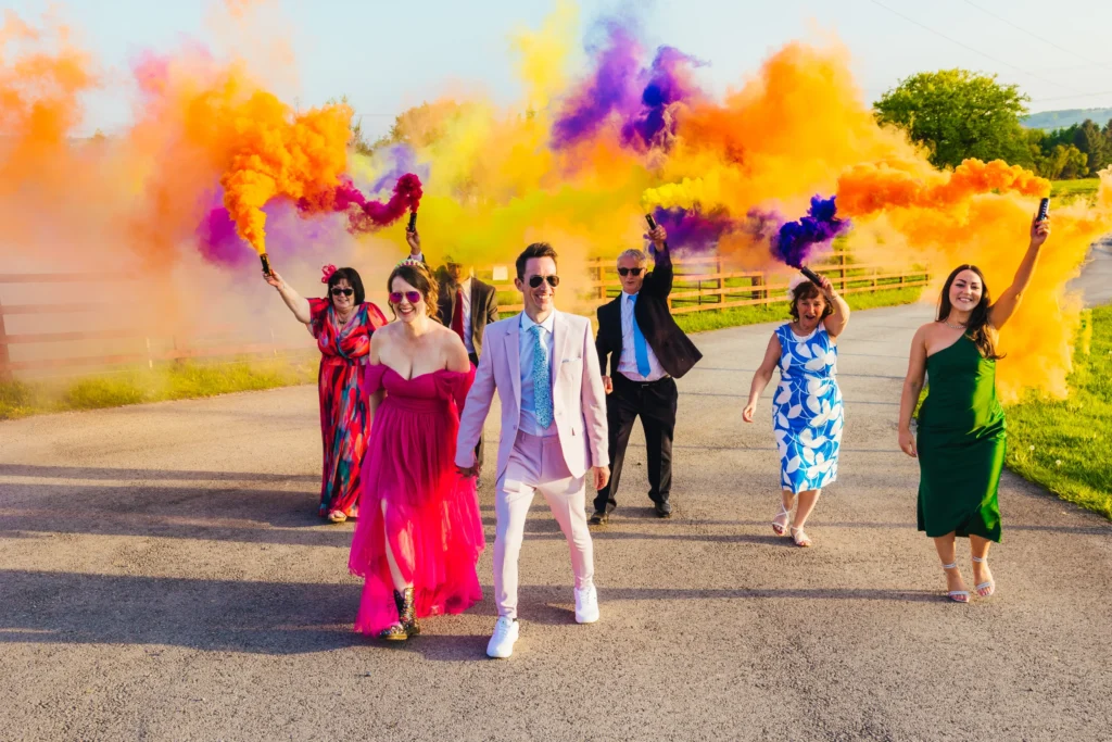 Couple walking both are wearing pink and they're being followed by a group of guests holding colourful smoke