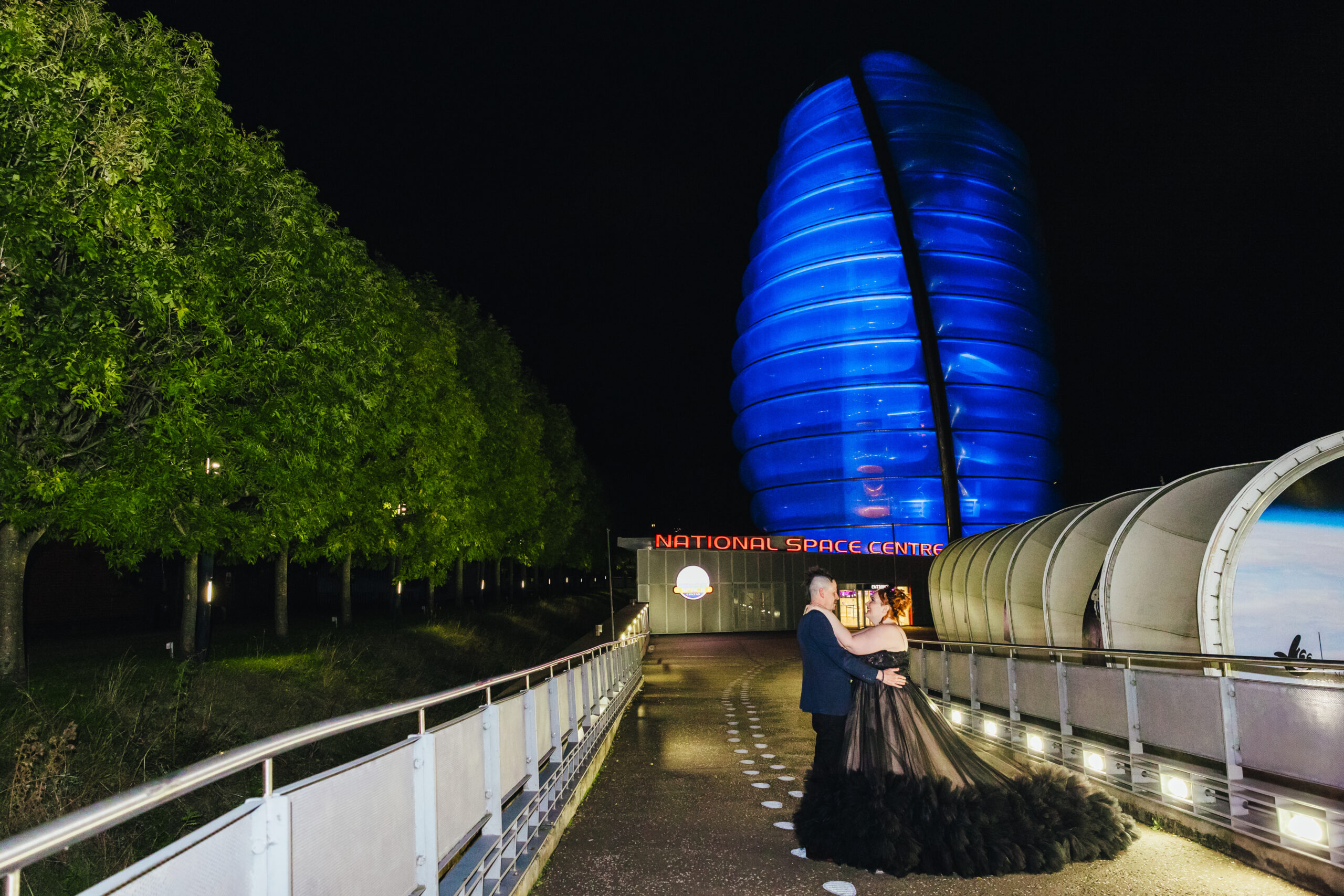 National Space Centre Wedding - Couple posing outside of National Space Centre in the dark it's lit up blue. The bride is wearing a black dress groom is in a suit.