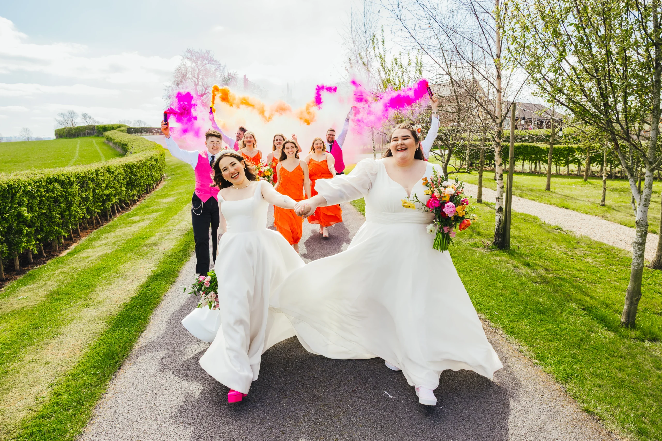 Couple swinging arms walking in front of their wedding party who are dancing and smiling holding colourful smoke. Both brides are holding beautiful colourful bouquets