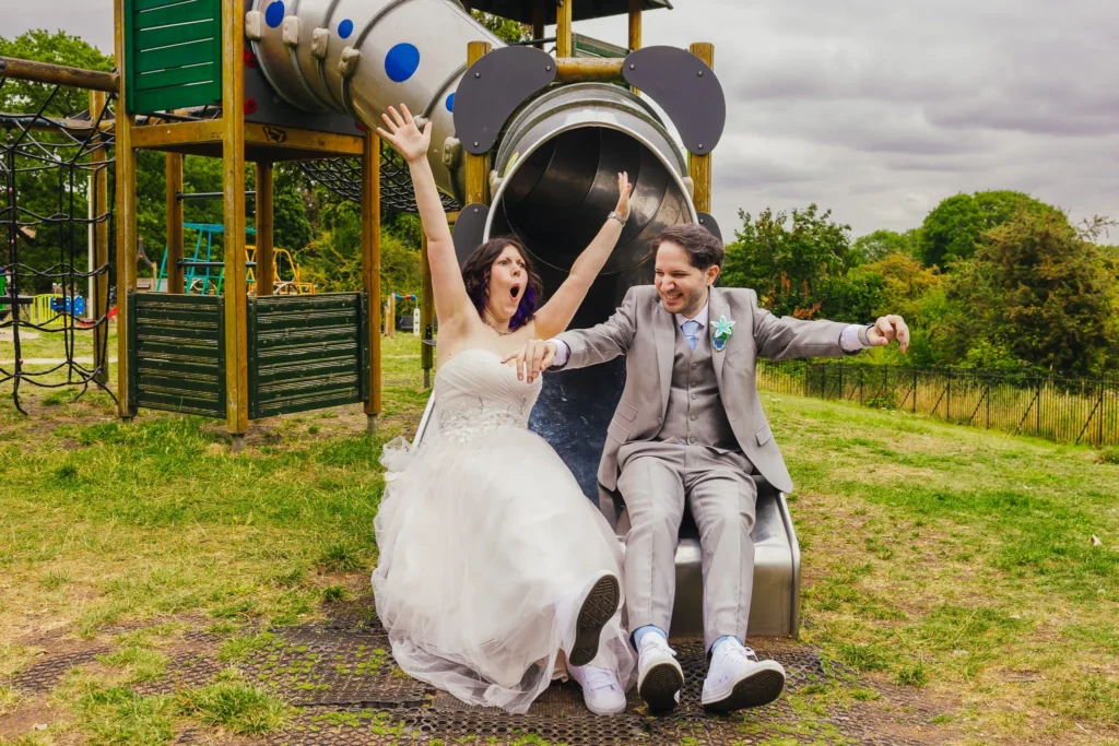 Bride and groom whooping as they come down a playground slide in their wedding outfits