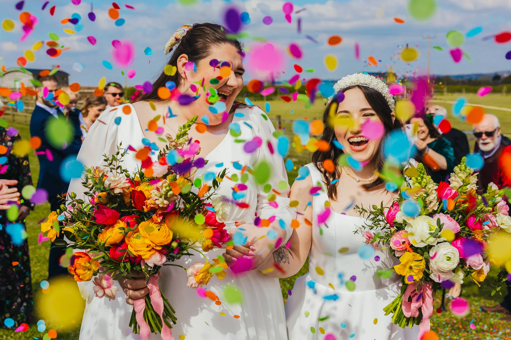 Two brides laughing as rainbow confetti flies around them, holding bright bouquets