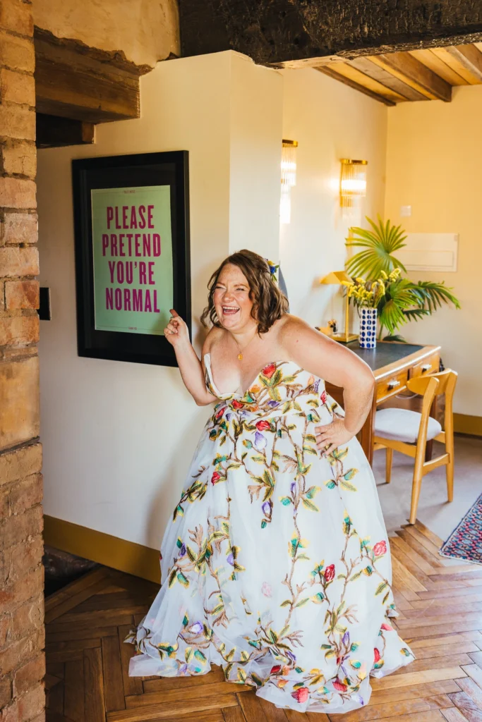 Bride in a colourful embroidered gown laughing and pointing at a Please Pretend You're Normal sign